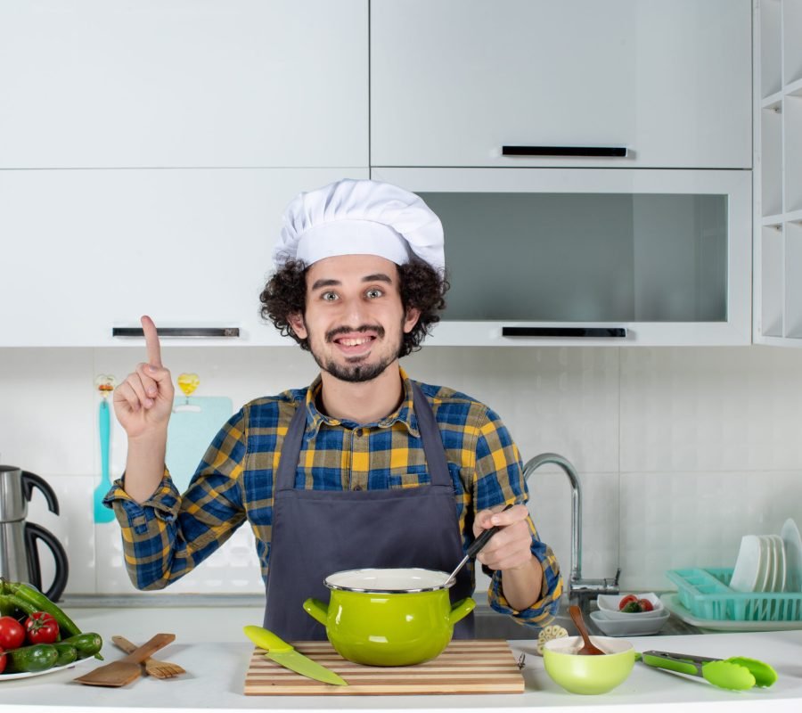 front-view-smiling-male-chef-with-fresh-vegetables-tasting-ready-meal-pointing-up-white-kitchen